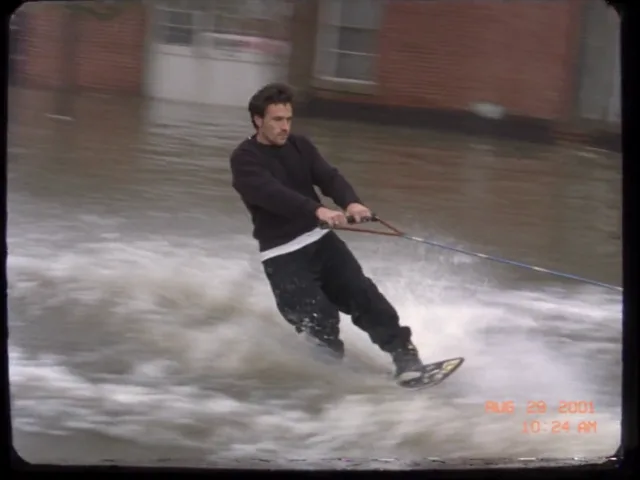 Ein Mann fährt Wakeboard durch eine überflutete Wohnstraße, hinter ihm zieht ein Motorboot zwischen halb versunkenen Autos