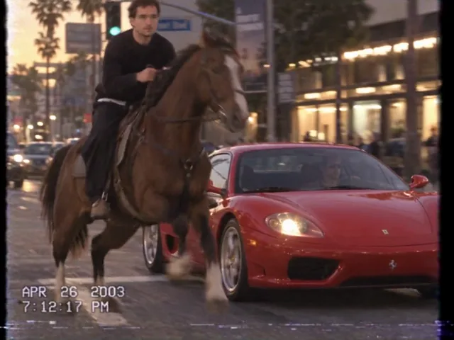 An einer Kreuzung im Abendlicht galoppiert ein Pferd mit Reiter neben einem roten Ferrari die Straße hinunter