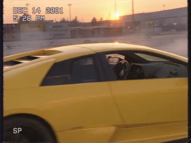Ein zweiter gelber Lamborghini zieht dicht an der Kamera vorbei, der Fahrer schaut aus dem Seitenfenster, hinter dem Wagen steht Rauch