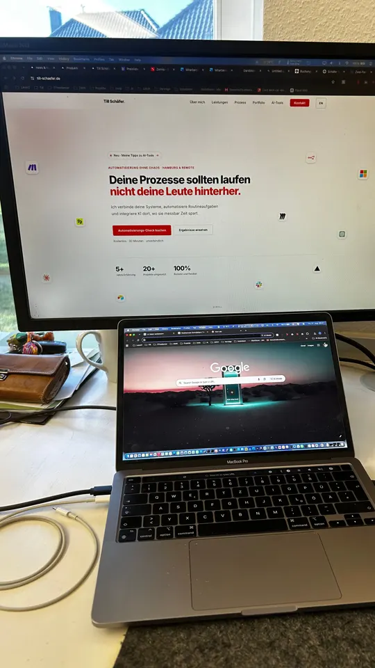 Der echte Schreibtisch: Monitor, aufgeklapptes MacBook, weiße Tischplatte, Fenster links
